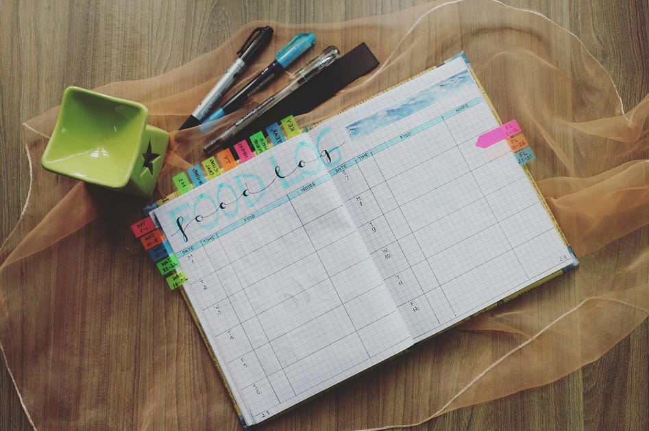 Open weekly planner on a wooden table, with colorful tab dividers, pens, and a green square pen holder nearby, plus a sheer fabric overlay.